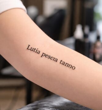 Braço de uma pessoa com uma tatuagem em latim no antebraço, em um estúdio de tatuagem com equipamentos ao fundo.