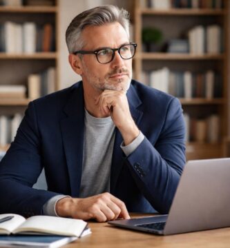 Homem de meia-idade sentado em uma mesa de escritório moderno, com expressão pensativa e roupas casuais elegantes.