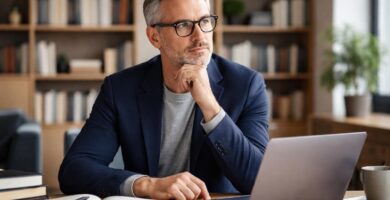 Homem de meia-idade sentado em uma mesa de escritório moderno, com expressão pensativa e roupas casuais elegantes.
